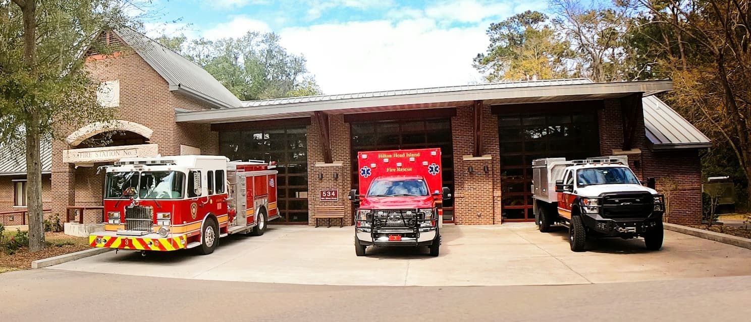 An engine, an ambulance and a utility truck parked on the apron in front of a station’s bay doors.
